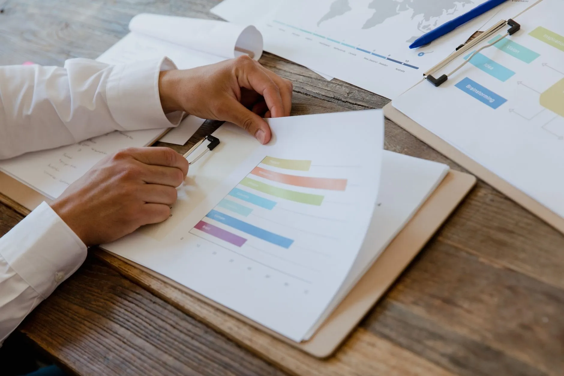 Hands turning a page of printed bar charts on a clipboard, more planning sheets spread over a wooden table