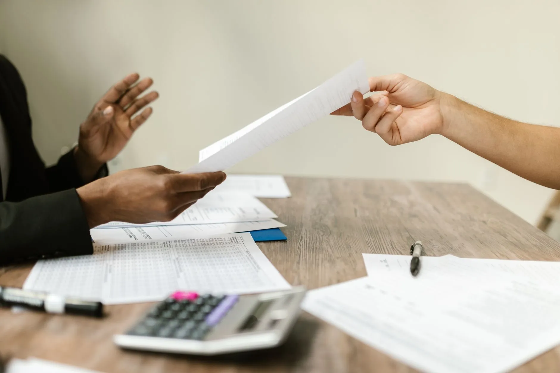 Hands passing a single printed page across a wooden table covered with forms and a calculator