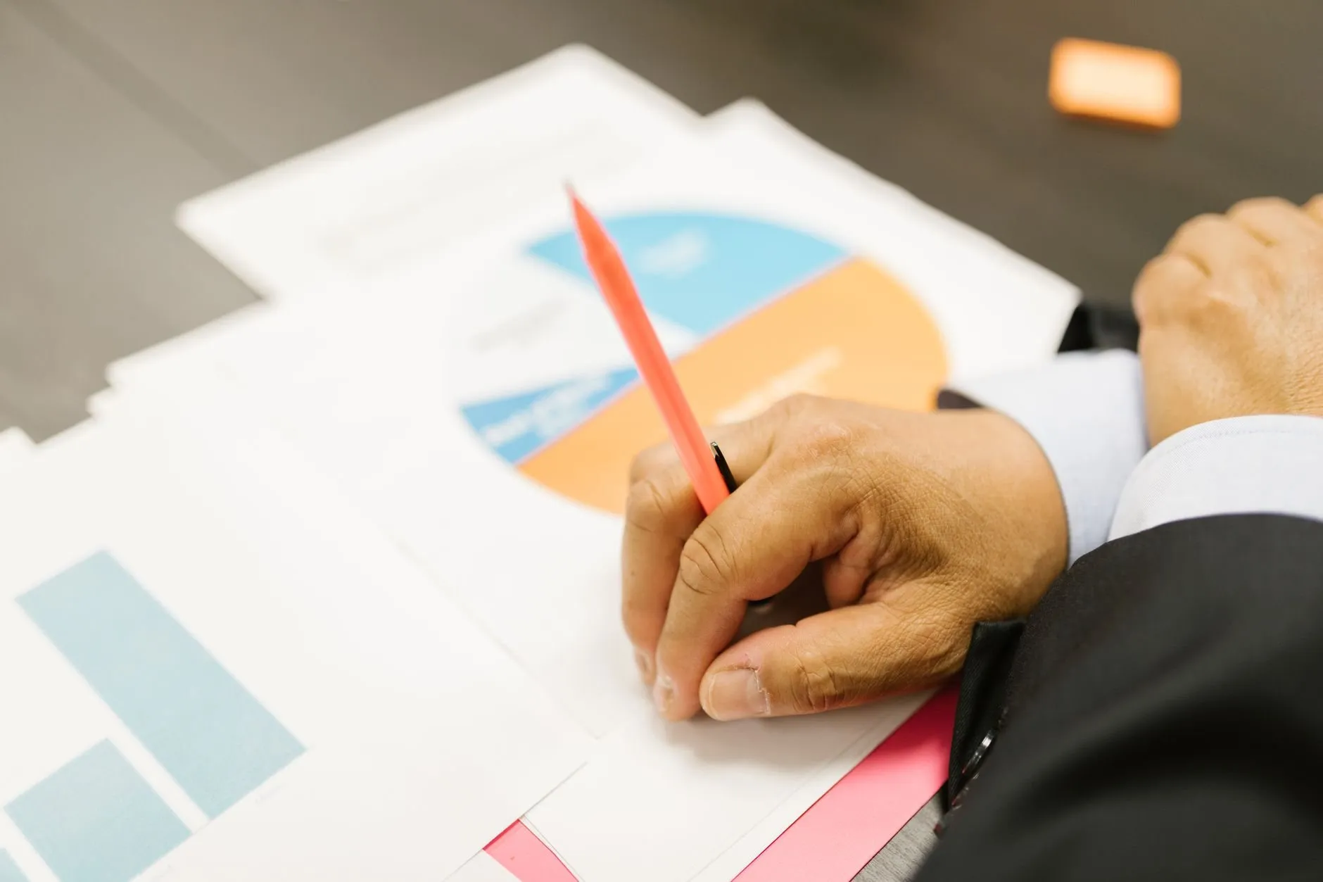 A hand writing on a printed pie chart with an orange pencil, other chart pages spread across the desk