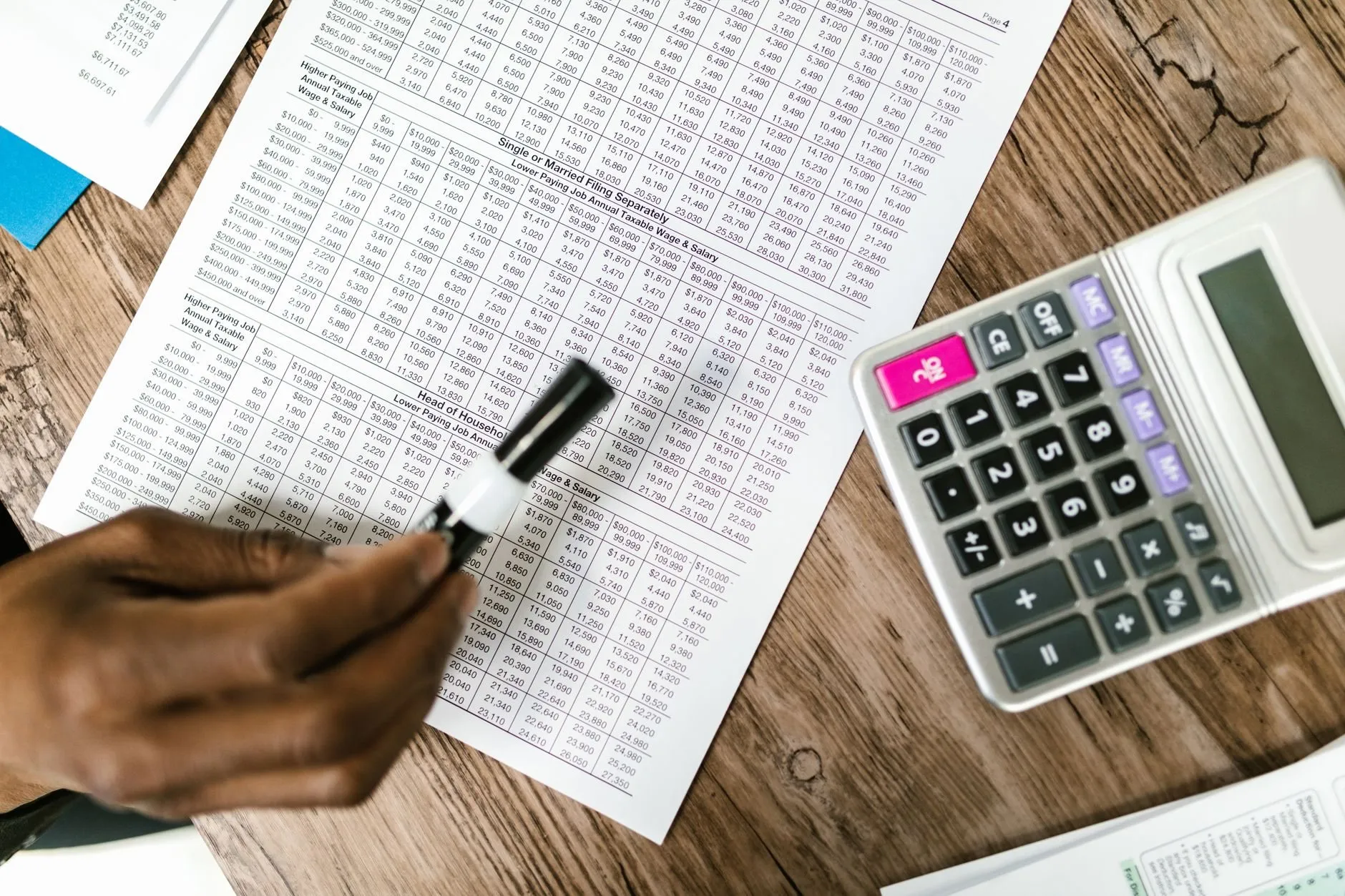 A hand holding a marker over a printed table of figures, a calculator beside it on a wooden desk
