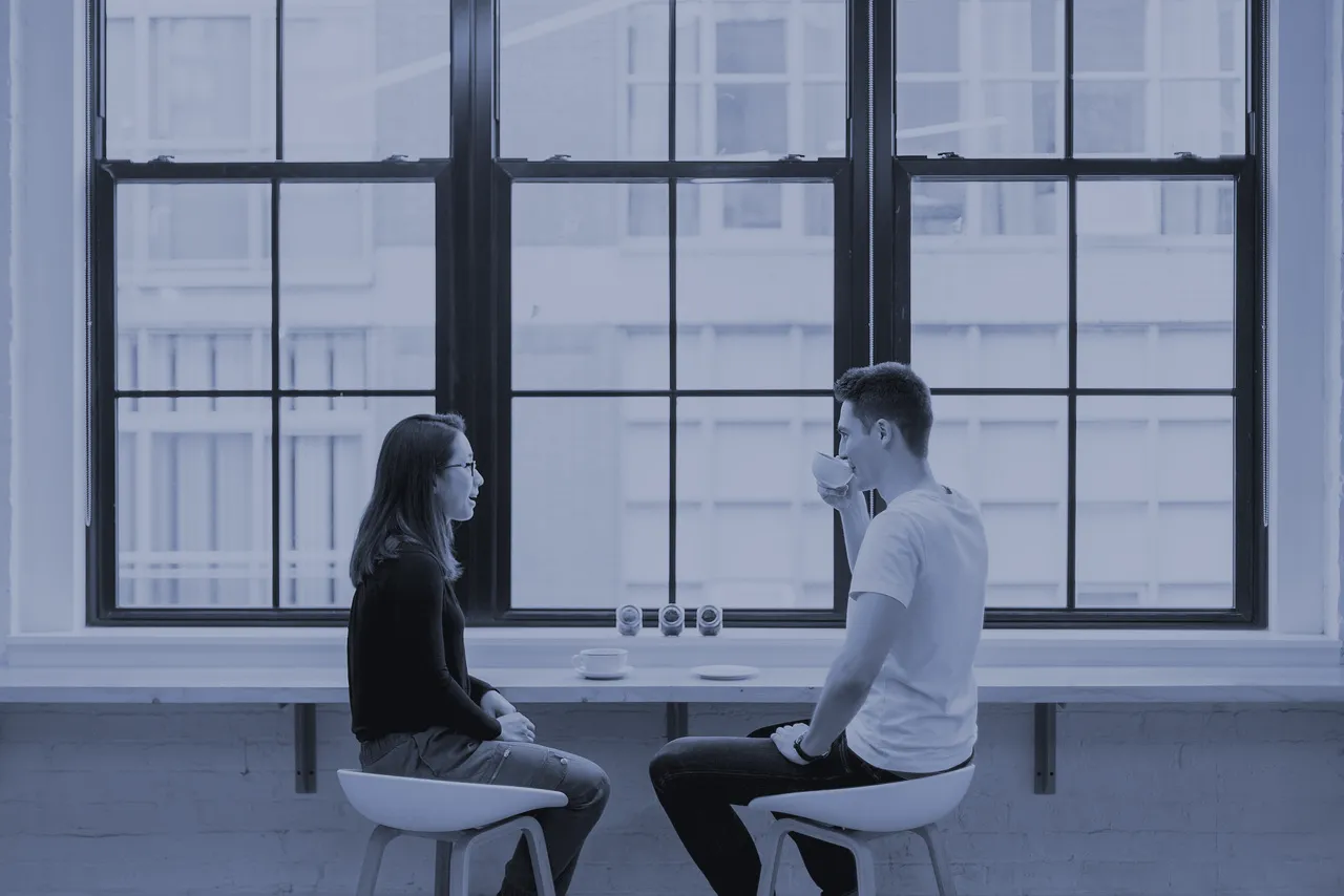 Two people talking across a counter at a tall gridded window.